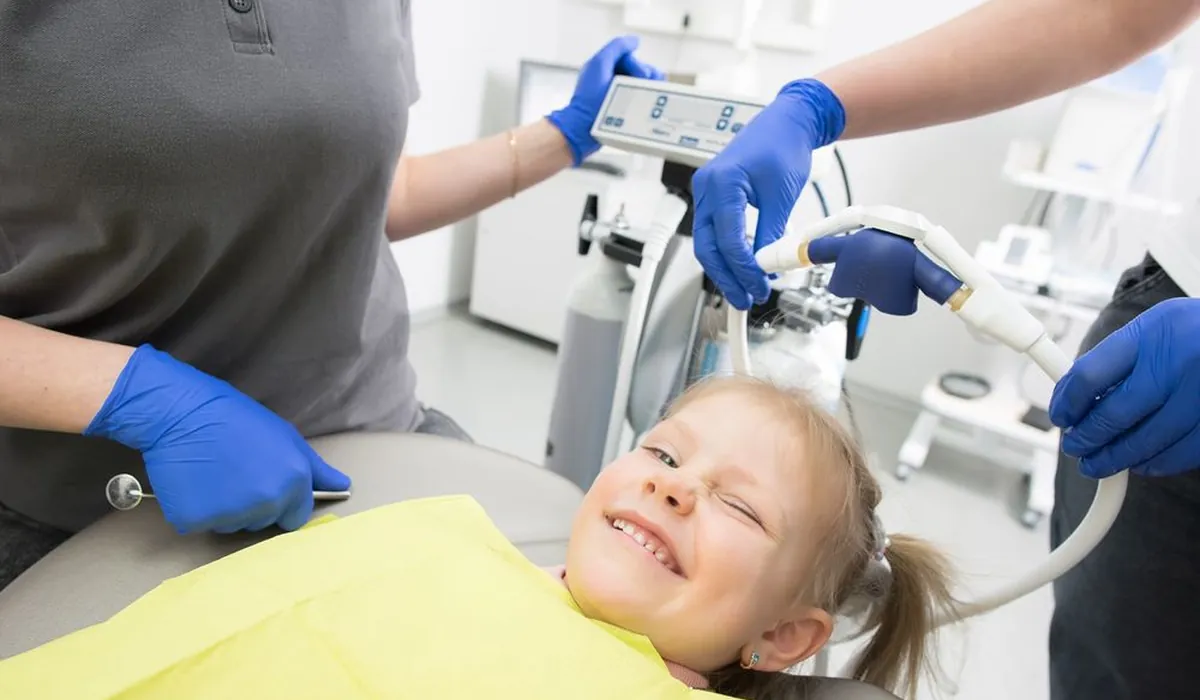 Diş koltuğunda sarı önlükle gülümseyen küçük kız ve klinik ekibi